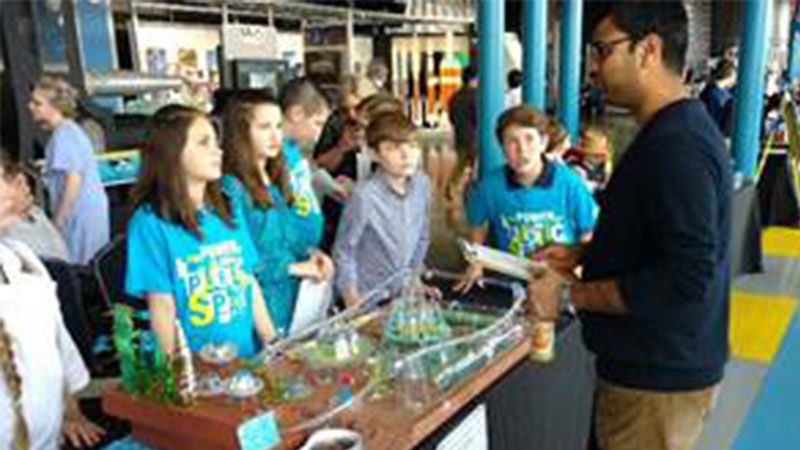 A man demonstrates a scientific experiment to a group of attentive children wearing matching blue shirts at a science fair.