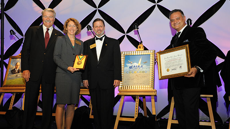 Four people posing with awards at a formal event, standing in front of a staged backdrop with geometric patterns and portraits.