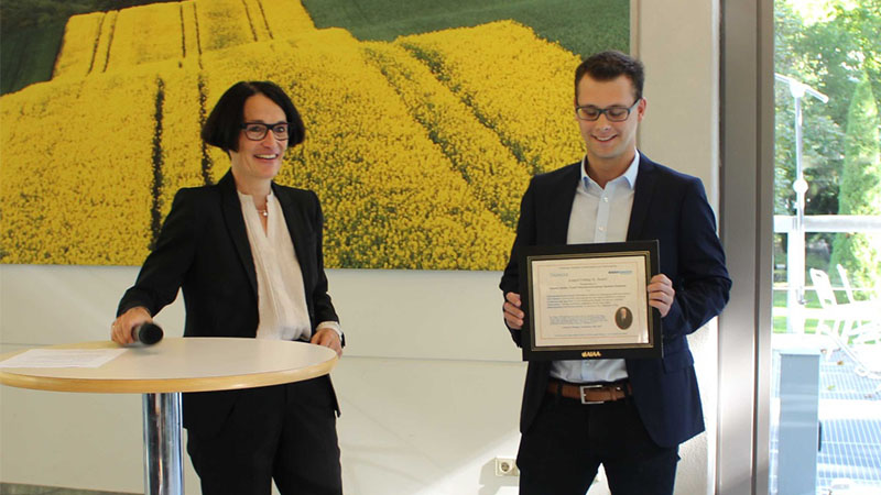 Two individuals, one holding a framed certificate, standing in front of a wall adorned with a large yellow field painting, in a well-lit room.