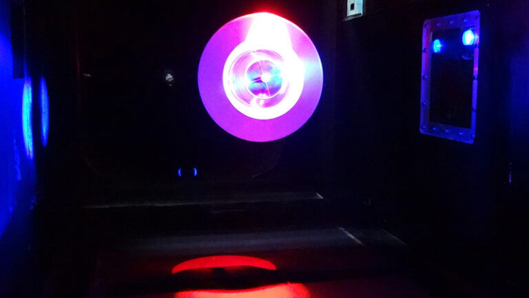 A neon-lit tunnel with a circular pink light at the end, reflecting on a glossy floor, surrounded by dark walls.