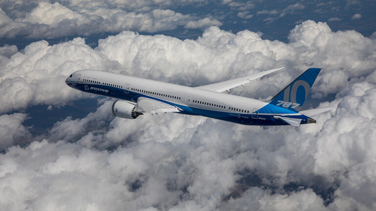 A boeing 787 aircraft flying above cloud cover, displaying boeing's livery on a clear day.