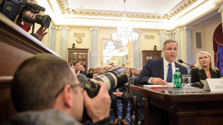 A man speaks at a hearing in an ornate room, surrounded by photographers and an attentive audience, including a woman seated next to him.