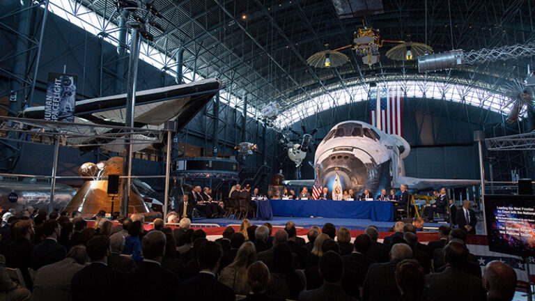 A formal gathering inside a hangar with a space shuttle and a large american flag in the background, attended by an audience in business attire.