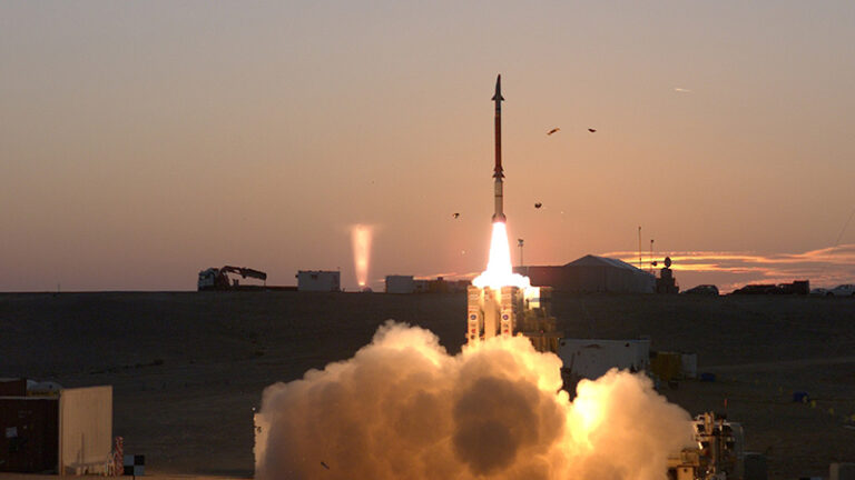 Rocket launching from a launch pad at dusk, with a bright flame and smoke trail, set against an orange sky.