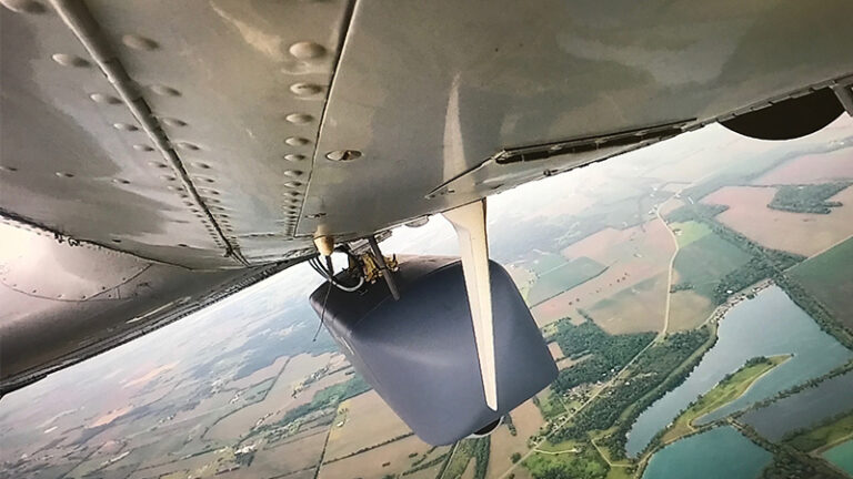 View from under an airplane wing showing a spinning propeller and a scenic aerial view of patchwork fields and lakes.