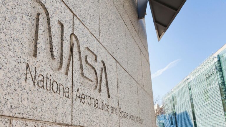 The NASA logo and "National Aeronautics and Space Administration" are etched on a stone wall with a background of modern buildings and a clear blue sky.