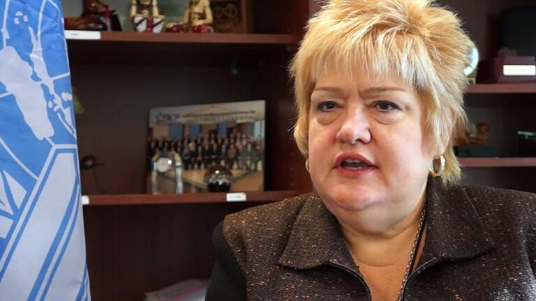 A woman with short blond hair is speaking. She is in an office setting with various items and a group photo on the shelf behind her.