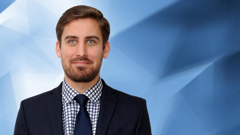 A man with short brown hair, beard, and mustache, dressed in a black suit, white and black checkered shirt, and navy blue tie, standing against a blue geometric background.
