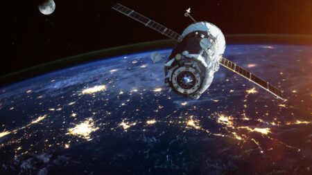 A satellite orbits Earth at night with city lights visible below; the moon is also seen in the background.