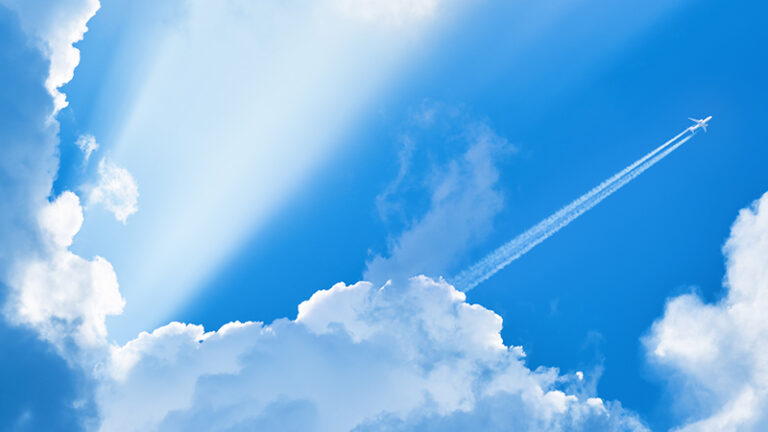 A jet airplane flies through a clear blue sky, leaving a trail of white contrails behind it. Sunrays penetrate through scattered, fluffy clouds.