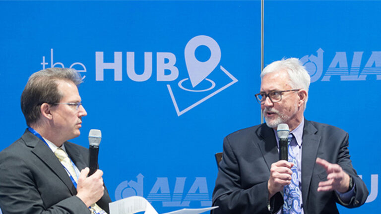 Two men in suits holding microphones are engaged in conversation in front of a blue backdrop with the words "the HUB" and "AIA.