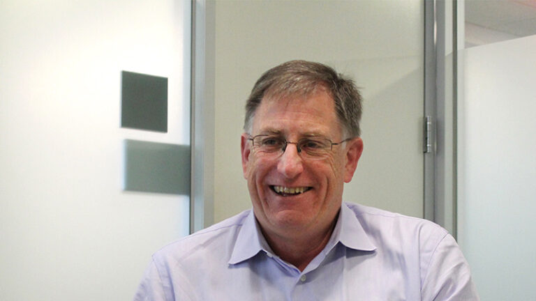 A man with glasses and a purple shirt is smiling, standing in an office with white walls and a glass panel door.