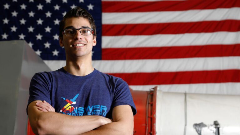 A person standing with crossed arms in front of a large American flag, wearing glasses and a T-shirt with colorful text and a graphic of an airplane.