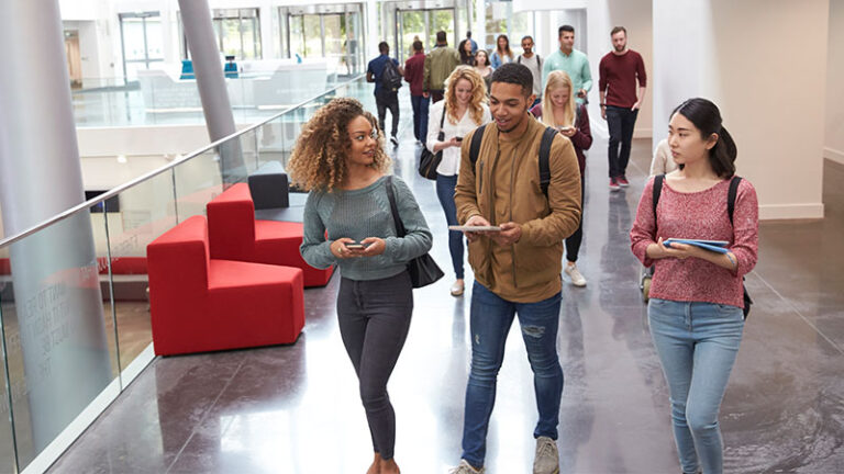 A group of students walks through a modern, well-lit building. Some are engaging with their phones and tablets, while others are chatting or walking in the background.