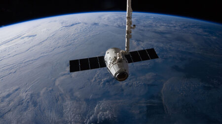 A satellite with solar panels orbits Earth with a mechanical arm attached, against the backdrop of the planet's blue and white cloud-covered surface.