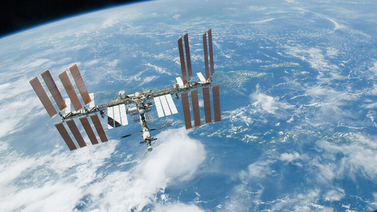 The International Space Station orbits above Earth, showing its solar panels extended with the planet's blue atmosphere and cloud-covered surface visible in the background.