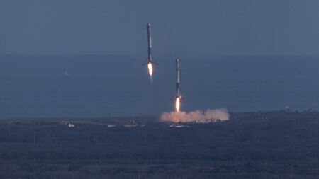 Two rockets are landing vertically on platforms near the coast. They have visible flames from their engines as they descend, with ocean and open land in the background.
