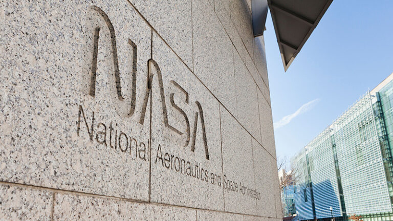 Exterior wall of a building with the NASA (National Aeronautics and Space Administration) logo and name engraved on it. A modern glass building is visible in the background.