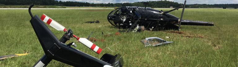A damaged helicopter lies in a grassy field, its tail section broken off and resting nearby. Debris from the crash is scattered around, and a runway is visible in the background.