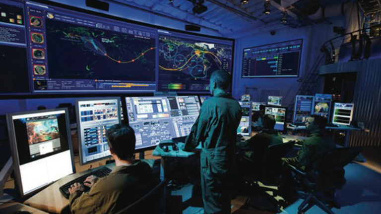 A group of people in a control room monitor various screens displaying maps, data, and graphics.