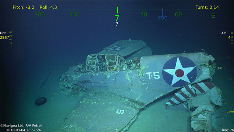 Underwater image of a sunken WWII airplane with visible USA markings, navigation data overlays, and the label "T-5" on the fuselage. Appears intact but shows some damage and marine growth.