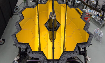 The James Webb Space Telescope with its large, gold hexagonal mirrors is being worked on by technicians in a laboratory setting.