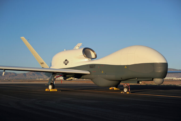 U.S. Navy drone aircraft parked on a runway during daytime with mountains in the background.