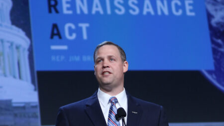 A man in a suit and tie speaks at a podium with a microphone. Behind him, a banner reads "RENAISSANCE ACT" and part of "REP. JIM." The background includes partial images of the U.S. Capitol and a globe.