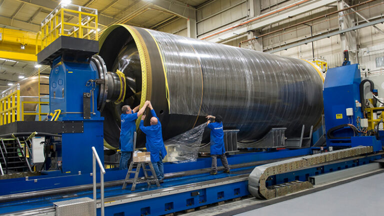 Three workers in blue uniforms inspect and work on a large industrial cylindrical machine in a manufacturing facility. The machine is silver with yellow accents and encased in plastic.