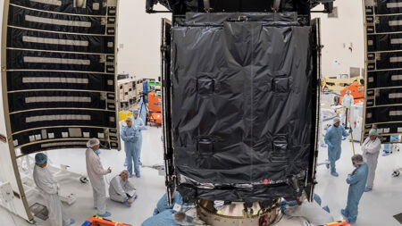 Several technicians in clean room suits work on a large, partially covered spacecraft in a laboratory setting.