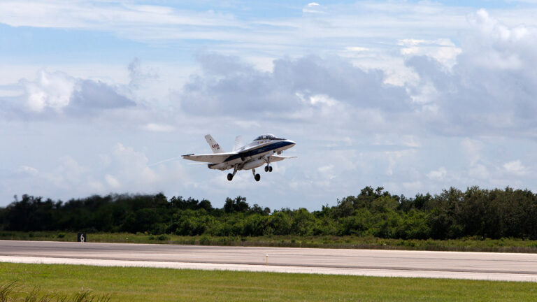 A small jet aircraft takes off from a runway with a cloud-filled sky in the background and greenery lining the sides of the runway.