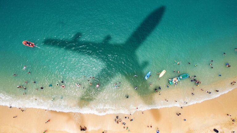 Aerial view of a beach with numerous people in the water and on the sand, with the large shadow of an airplane cast over the water and part of the beach.