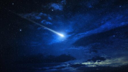 A bright meteor streaks across a star-filled night sky, illuminating the surrounding clouds with a vivid blue glow.
