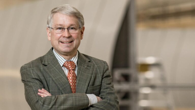 A man with gray hair and glasses stands indoors, wearing a green checked suit, striped shirt, and orange patterned tie, with arms crossed and smiling. Blurred background includes industrial elements.