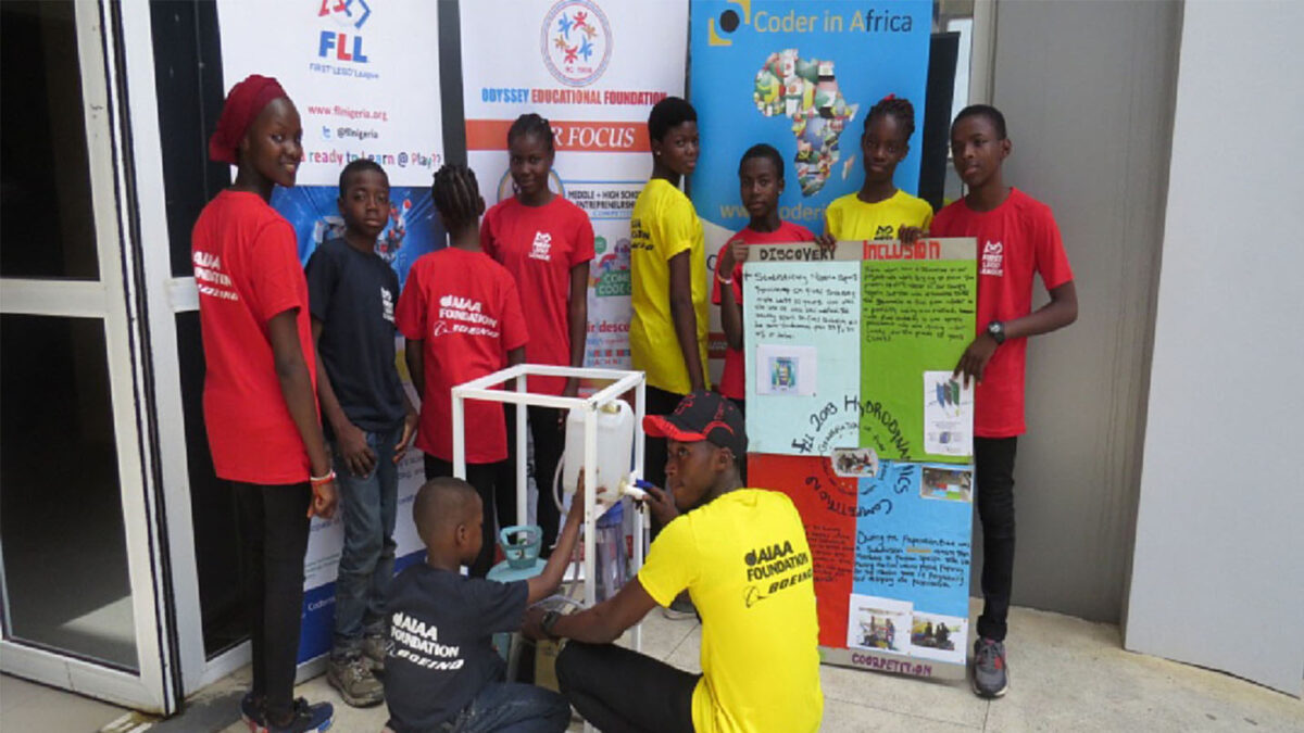 A group of young people in brightly colored shirts stand around a display of posters and a small machine, in front of educational organization banners.