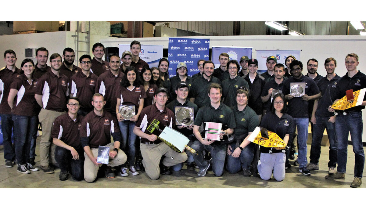 A large group of people poses for a photo in a warehouse-like space, some holding awards and colorful model airplanes. They wear matching maroon shirts.