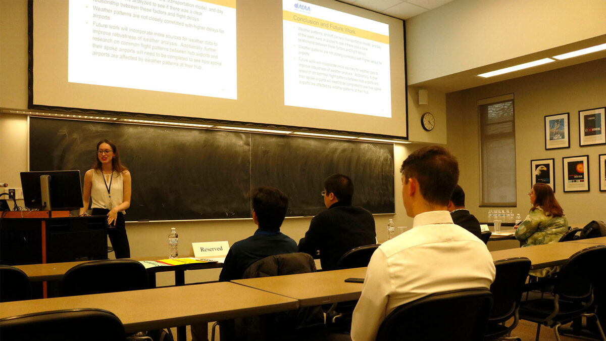 A person is giving a presentation to a small audience in a classroom. The presentation slides are projected onto a screen at the front of the room. Several people are seated and listening attentively.