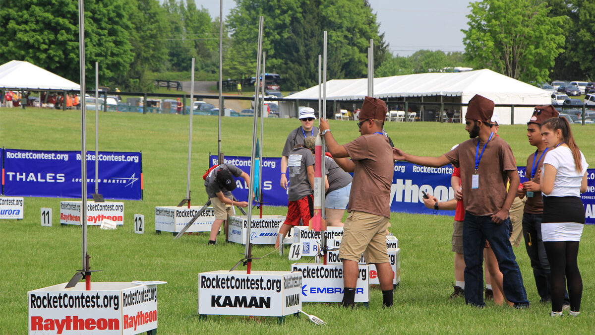 Participants prepare their rockets for launch during a rocket contest on a grassy field, with tents and sponsors' banners in the background.