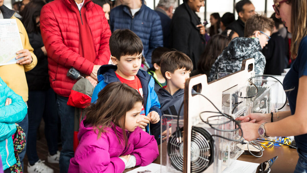 Children and adults observe and interact with science exhibits at a crowded indoor event.