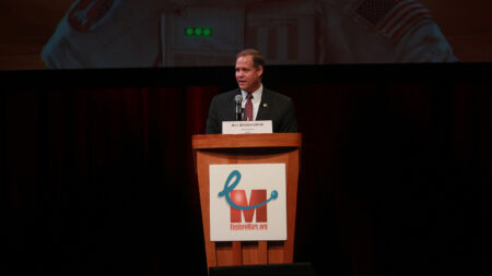 A person in a suit speaks at a podium labeled "exploremars.org." The name plaque on the podium reads "Jim Bridenstine." There is a backdrop of an astronaut suit behind the speaker.