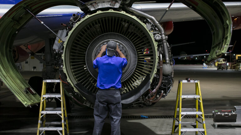 A technician wearing a blue shirt and cap works on the large exposed engine of an airplane at night. Two ladders are positioned on either side of the engine.