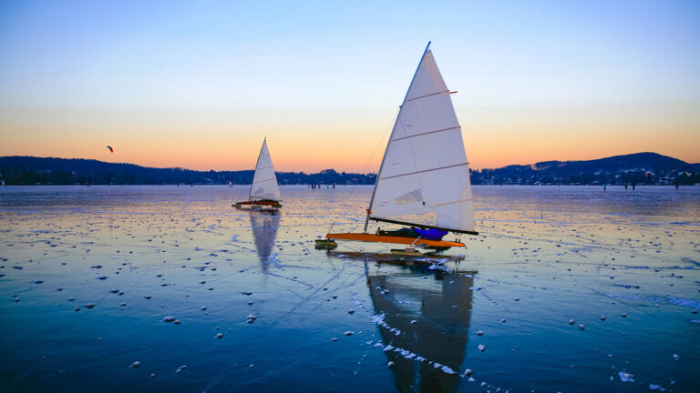 Two iceboats with white sails glide across a frozen lake during sunset, with hills and buildings in the background.