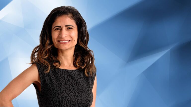 A woman with long dark hair, wearing a sleeveless black patterned dress, stands in front of a blue geometric background.