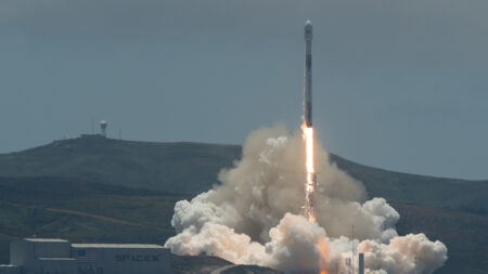 A SpaceX Falcon 9 rocket launches from a base, emitting flames and smoke while ascending into a cloudy sky over a hilly landscape.