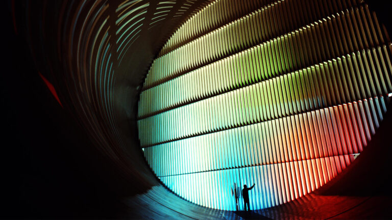 A person stands with arms raised inside a large, curved structure illuminated by colorful lights, with vertical slats casting shadows.