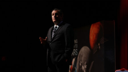 A man in a suit gestures as he stands on a stage in low lighting, with a backdrop featuring images of Mars and space-related themes.