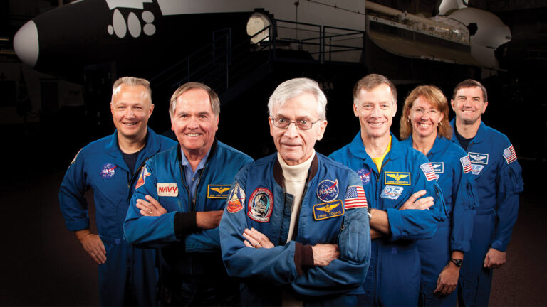 Group of six people wearing NASA uniforms stand together, smiling. A space shuttle cockpit is visible in the background.