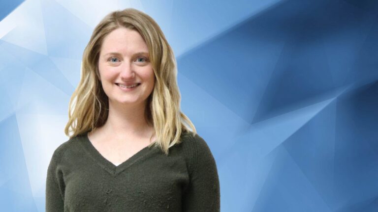A person with long blond hair wearing a dark green sweater stands against a blue geometric background, smiling at the camera.