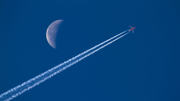 A red airplane flies across the sky, leaving a contrail behind, with a visible crescent moon in the background.