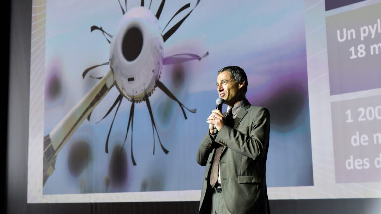 A man in a suit is speaking into a microphone on stage in front of a large screen displaying an image of a futuristic device with projecting appendages.